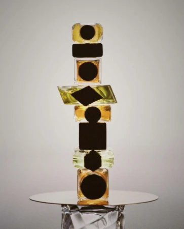Tower of geometric-shaped glass jars on a reflective surface, featuring clear and yellow liquids and abstract black labels.