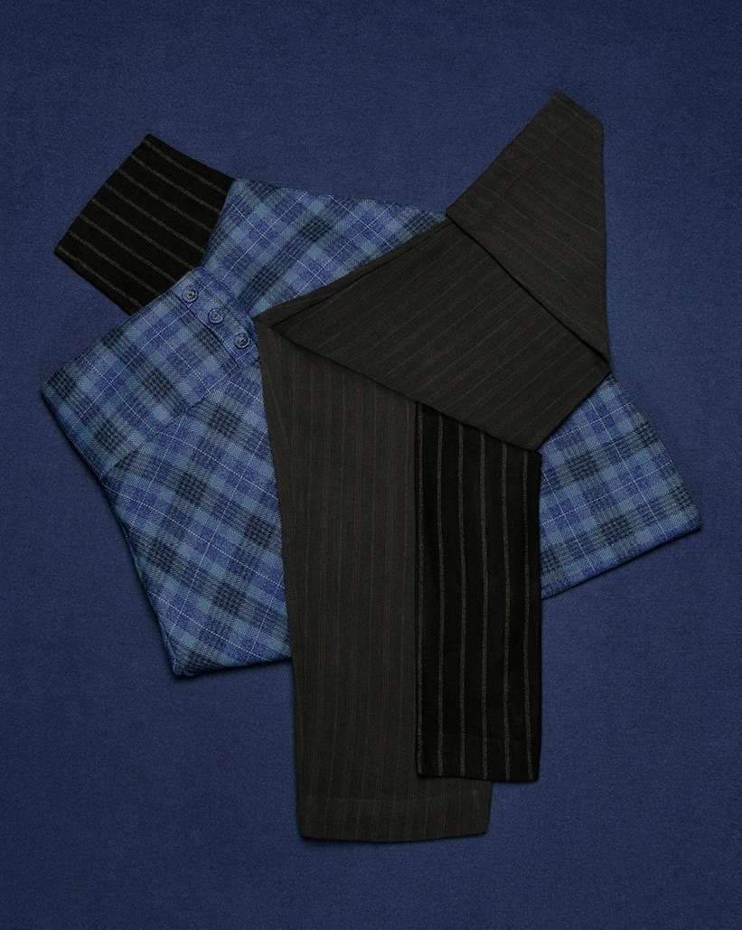 Still life of folded tailored garments including a blue plaid button-down shirt and dark pinstripe wool pants over a navy blue background.