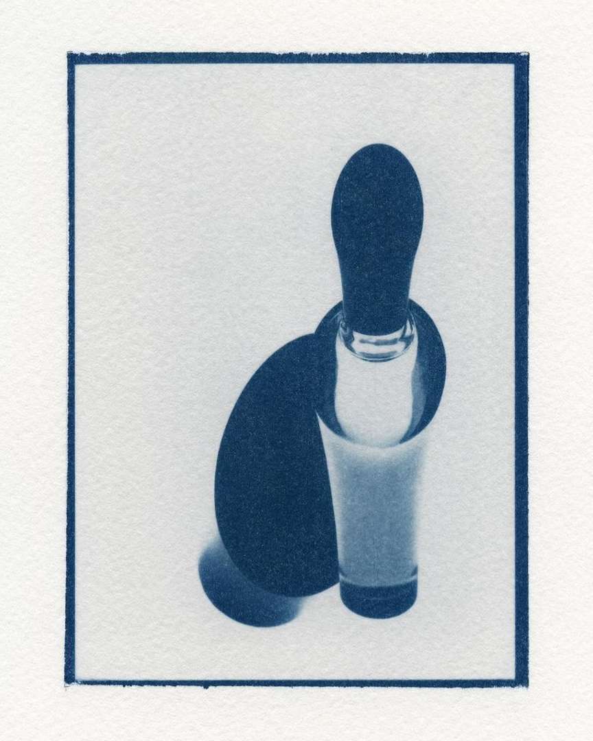 Cyanotype photograph of a vintage glass bottle resting in a transparent glass with a solid dark shadow against a white backdrop.