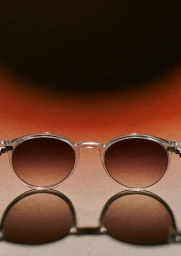 Close-up of translucent round sunglasses resting on a reflective surface with a warm gradient background.