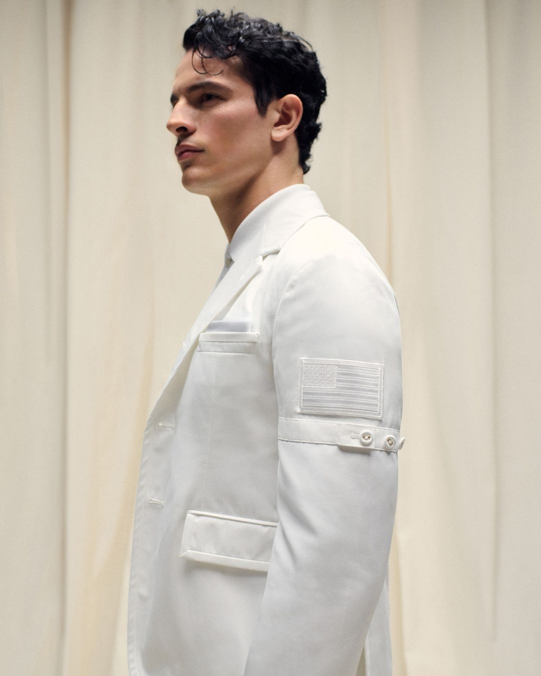 Adult male in profile wears a white jacket with a US flag patch in an indoor setting. Fashion-forward and patriotic.