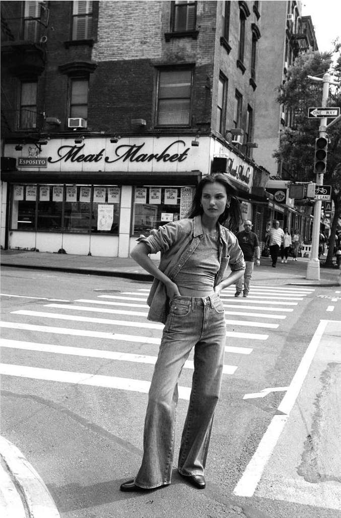 A woman in denim stands on a crosswalk in front of a brick building with a 'Meat Market' sign in a black and white fashion shot.