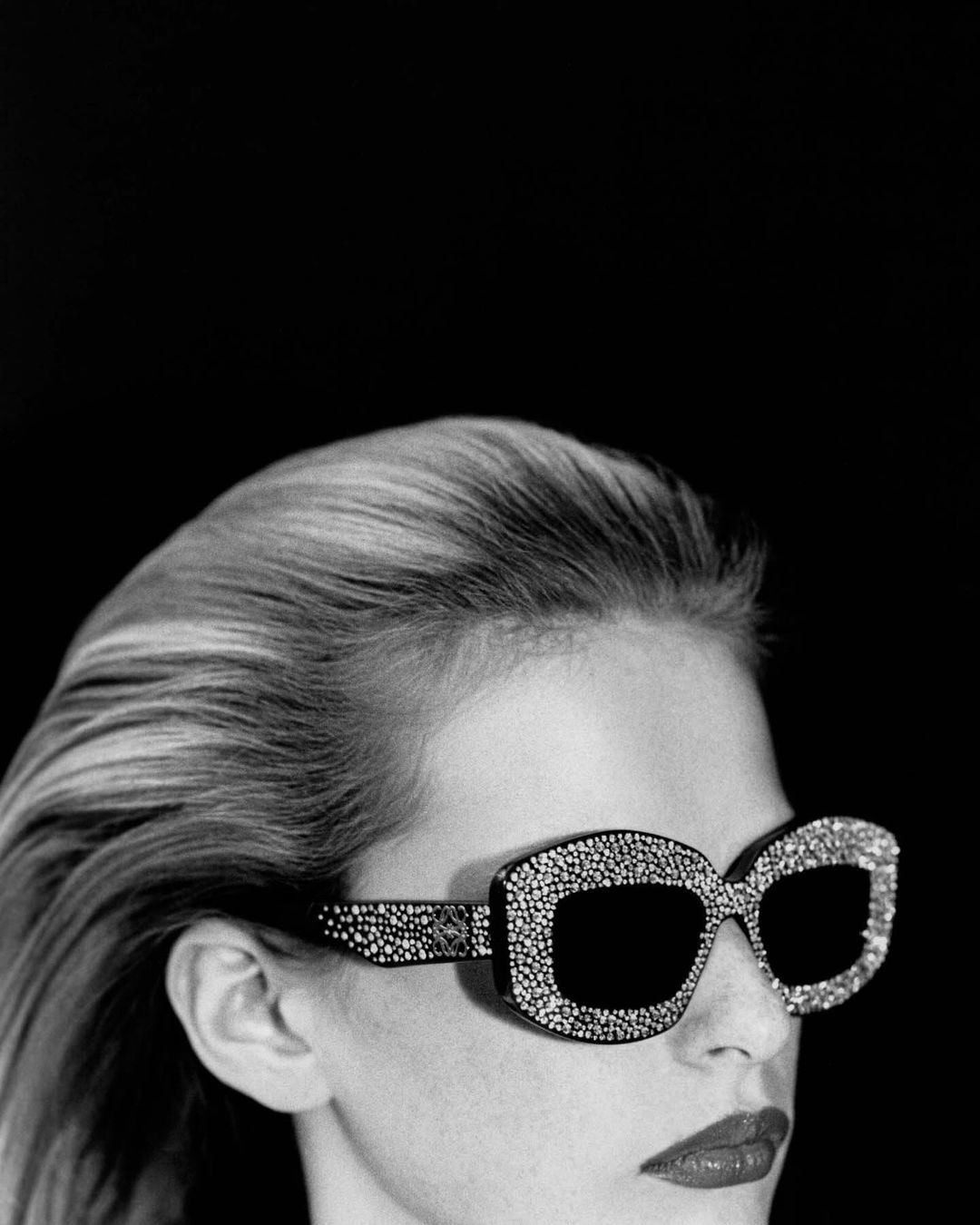 Black and white portrait of a woman with blonde hair wearing large, jewel-encrusted sunglasses against a plain black background.