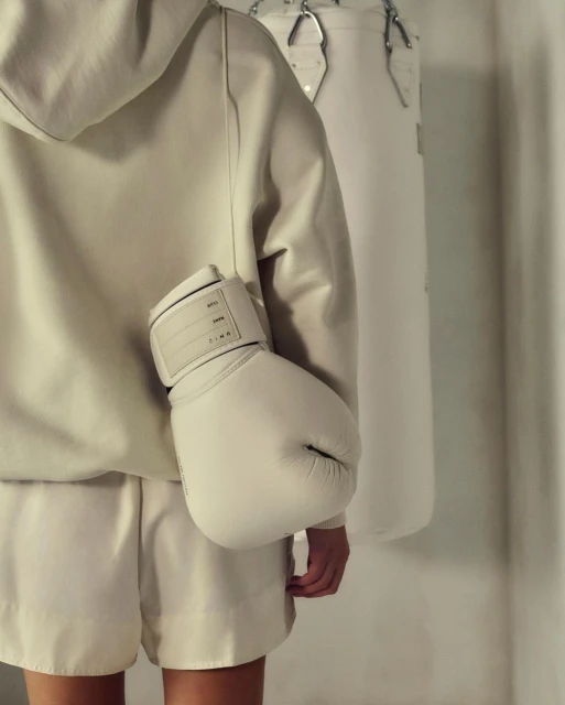 Young adult in cream-colored hoodie and shorts, holding boxing glove, posing in front of a white punching bag indoors.