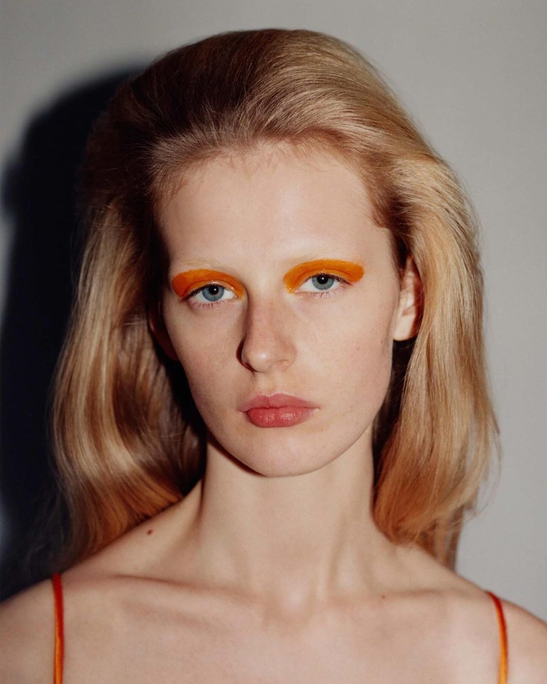 Close-up portrait of a blonde adult woman with bright orange eye makeup, posing in front of a neutral grey background.