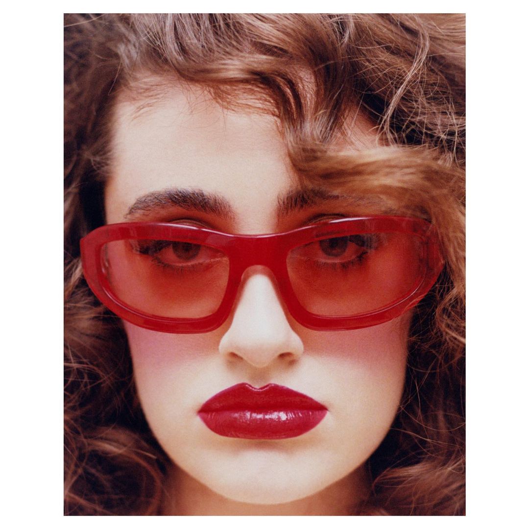 Close-up portrait of a brunette adult woman with dark eyes wearing red lipstick and red sunglasses with a white background.
