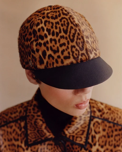 A woman wears a striking leopard-print hat and jacket with a black turtleneck. The animal print fashion and neutral backdrop highlight her style.