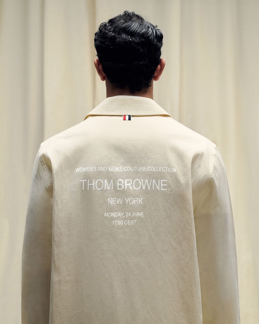 Back view of a light beige jacket with a small red, white, and blue tag on the collar, and 'Thom Browne' and 'New York' text on the back.
