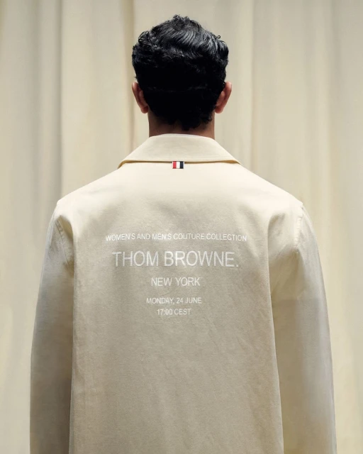 Back view of a light beige jacket with a small red, white, and blue tag on the collar, and 'Thom Browne' and 'New York' text on the back.
