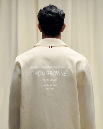 Back view of a light beige jacket with a small red, white, and blue tag on the collar, and 'Thom Browne' and 'New York' text on the back.