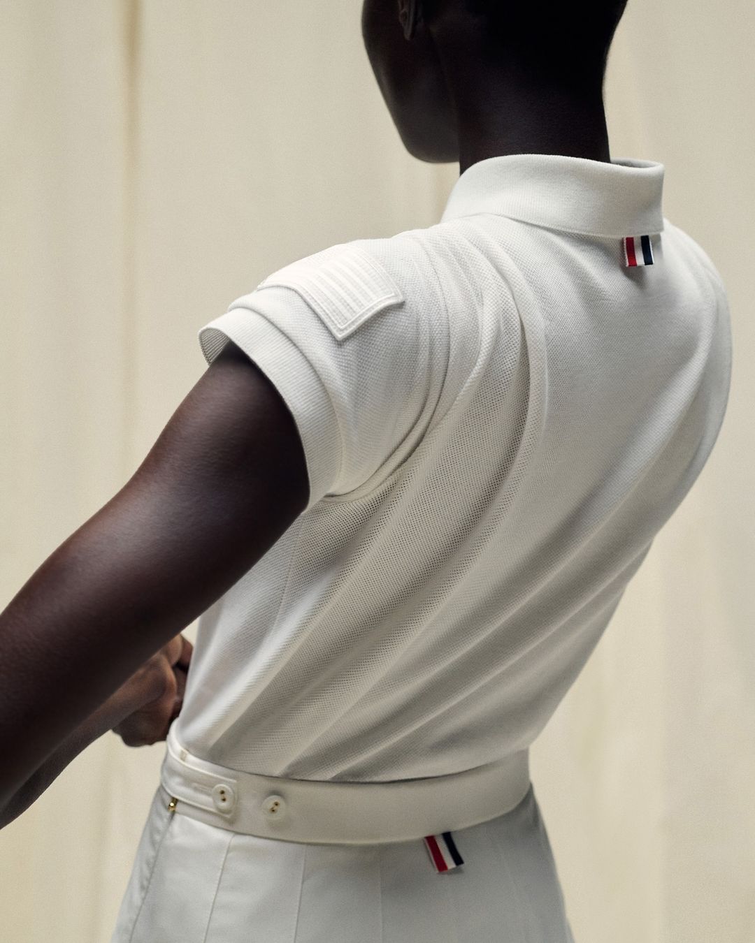 Dark-skinned model wearing a white polo shirt and skirt with red, white, and blue accents, stands against an off-white background in a fashion shot.