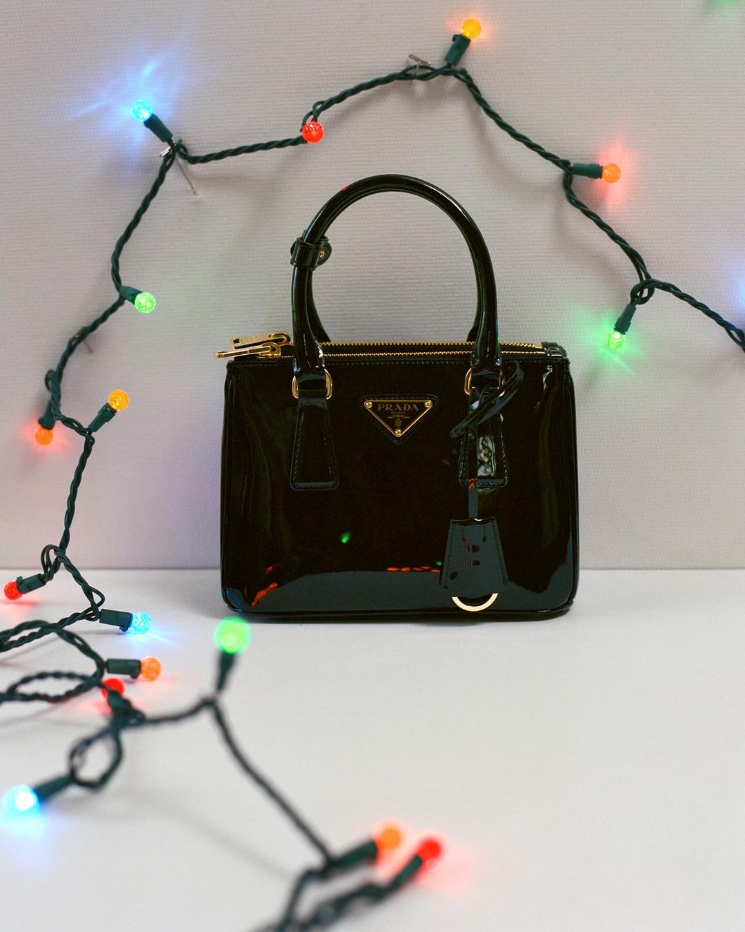 Glossy black Prada handbag sits in front of a string of festive multicolored Christmas lights on a white surface in a studio setting.