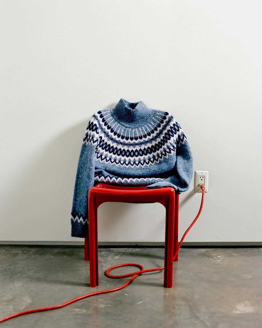Blue patterned sweater on a red plastic chair indoors with a red electrical cord plugged into the wall, in a studio setting.