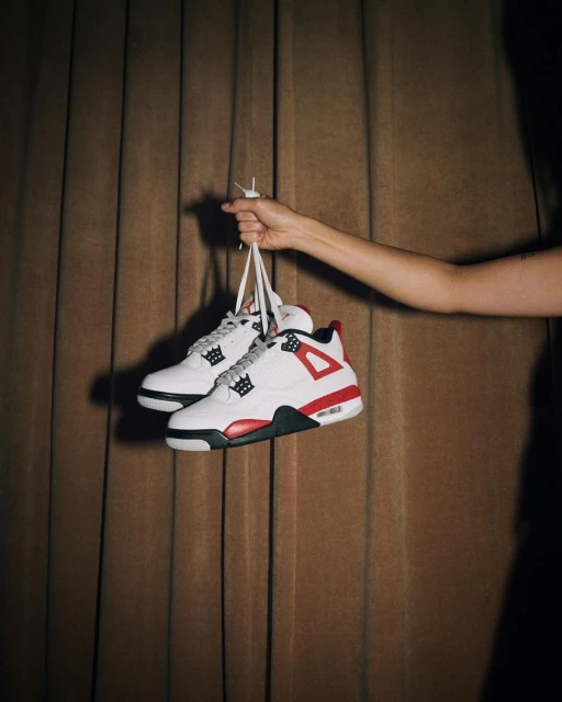 White and red Air Jordan 4 sneakers hanging by the laces from a person's hand against a brown curtain backdrop.