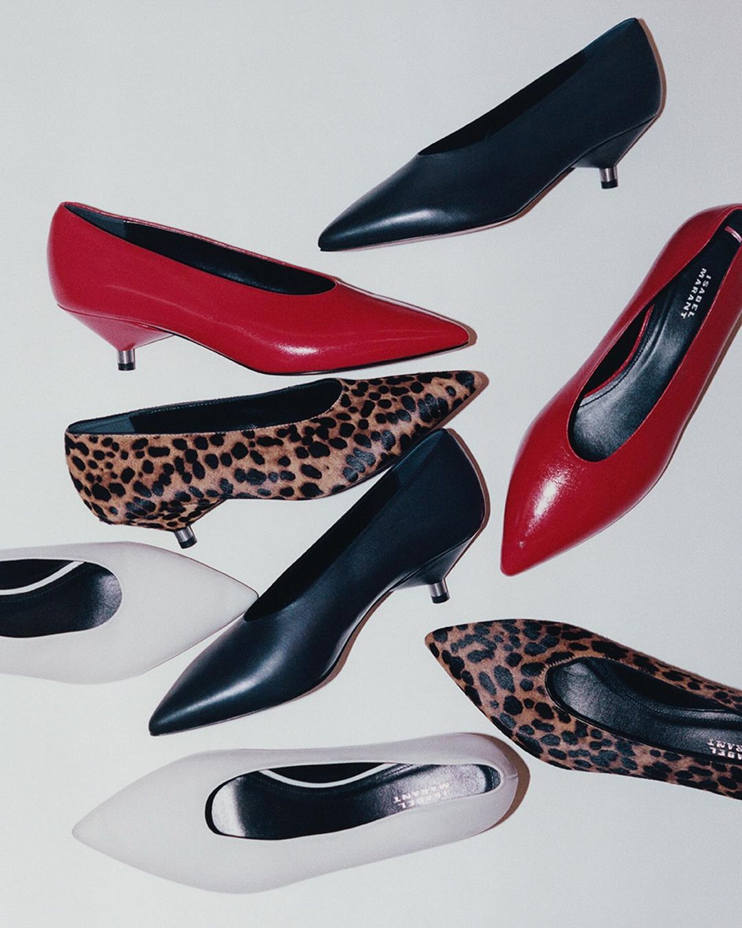 Collection of kitten heel pumps in red, white, leopard print and black. Fashionable women's shoes with pointed toes are scattered on a white surface.