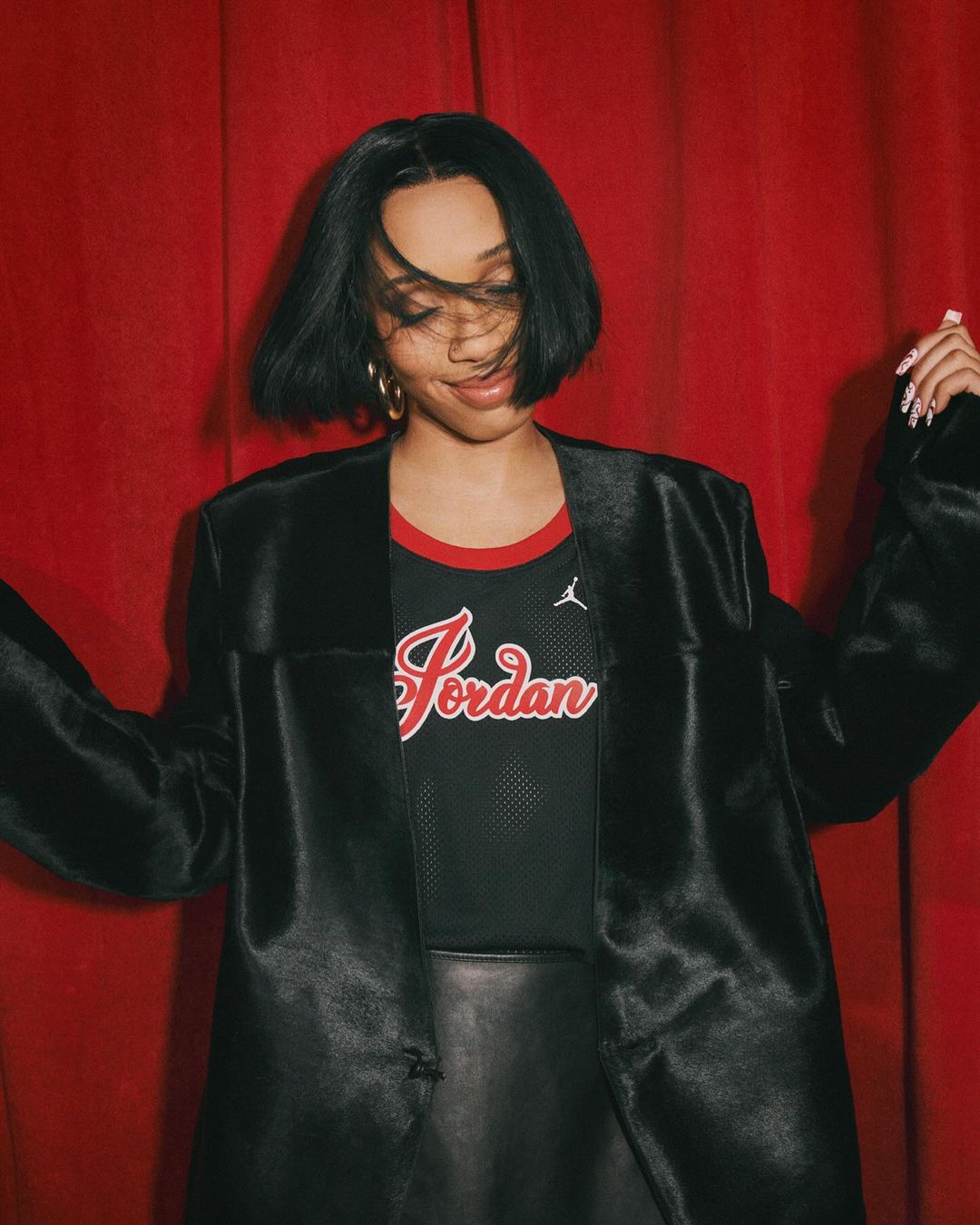 Portrait of an adult woman with a black silky jacket and black Jordan t-shirt posing in front of a red curtain in a studio.