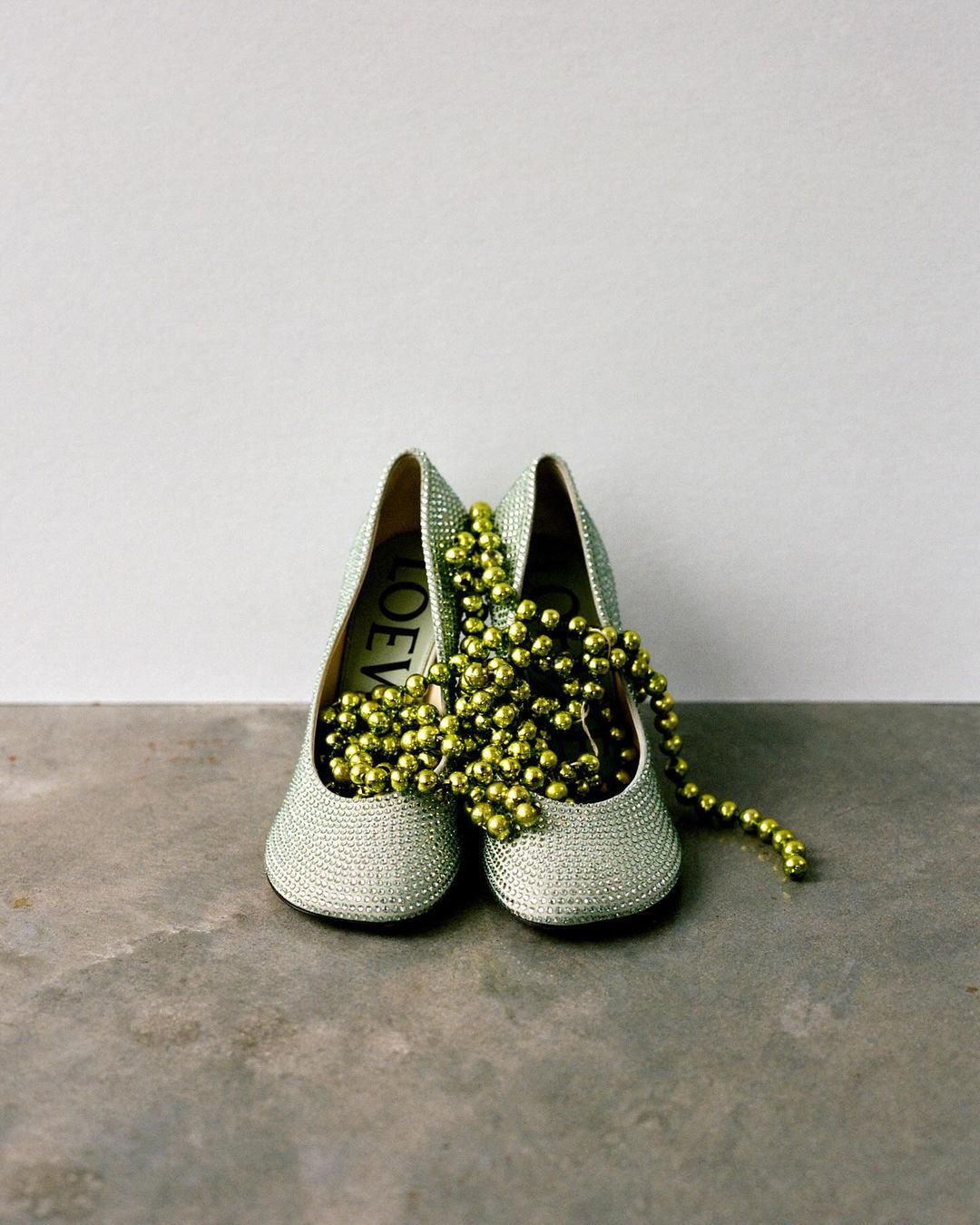 Still life studio shot of a pair of silver-toned heeled shoes with a string of gold beads draped over them, set against a plain backdrop.