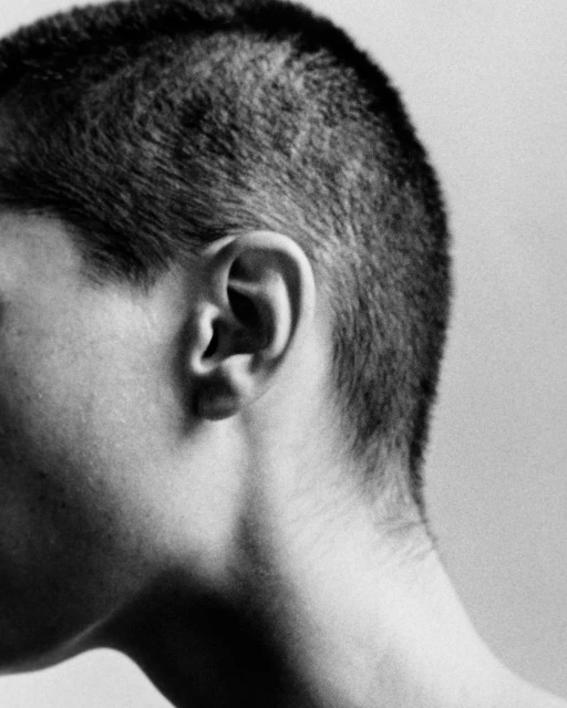 Monochrome portrait of an adult with a very short haircut, featuring the ear, neck, and hairline in a minimalist composition.