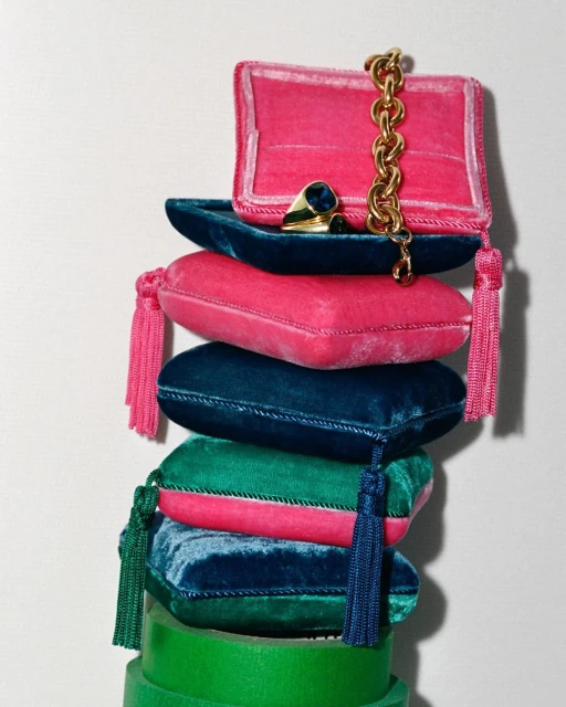 Stacked velvet jewelry pillows in pink, teal, and green with gold chain and ring, studio lighting, and bright colors.