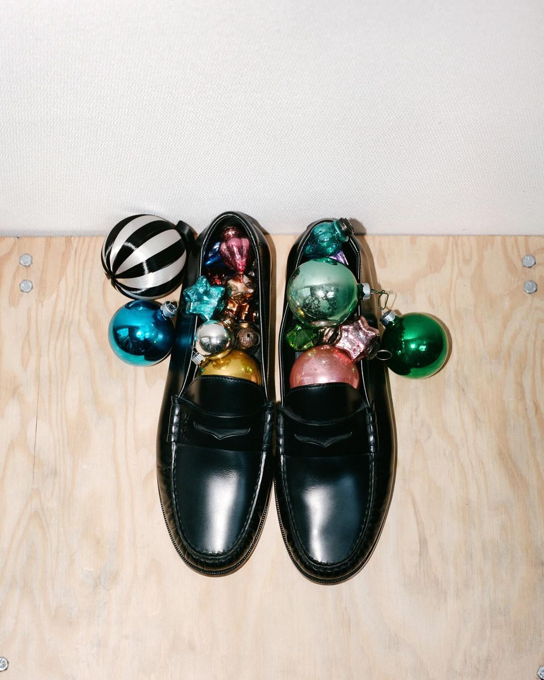 Pair of black leather loafers filled with colorful Christmas ornaments on a light wood surface against a plain white background.