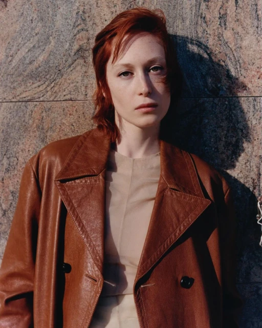 Portrait of a red-haired woman wearing a brown leather coat in front of a granite wall with a shadow. Fashion shot with natural light.