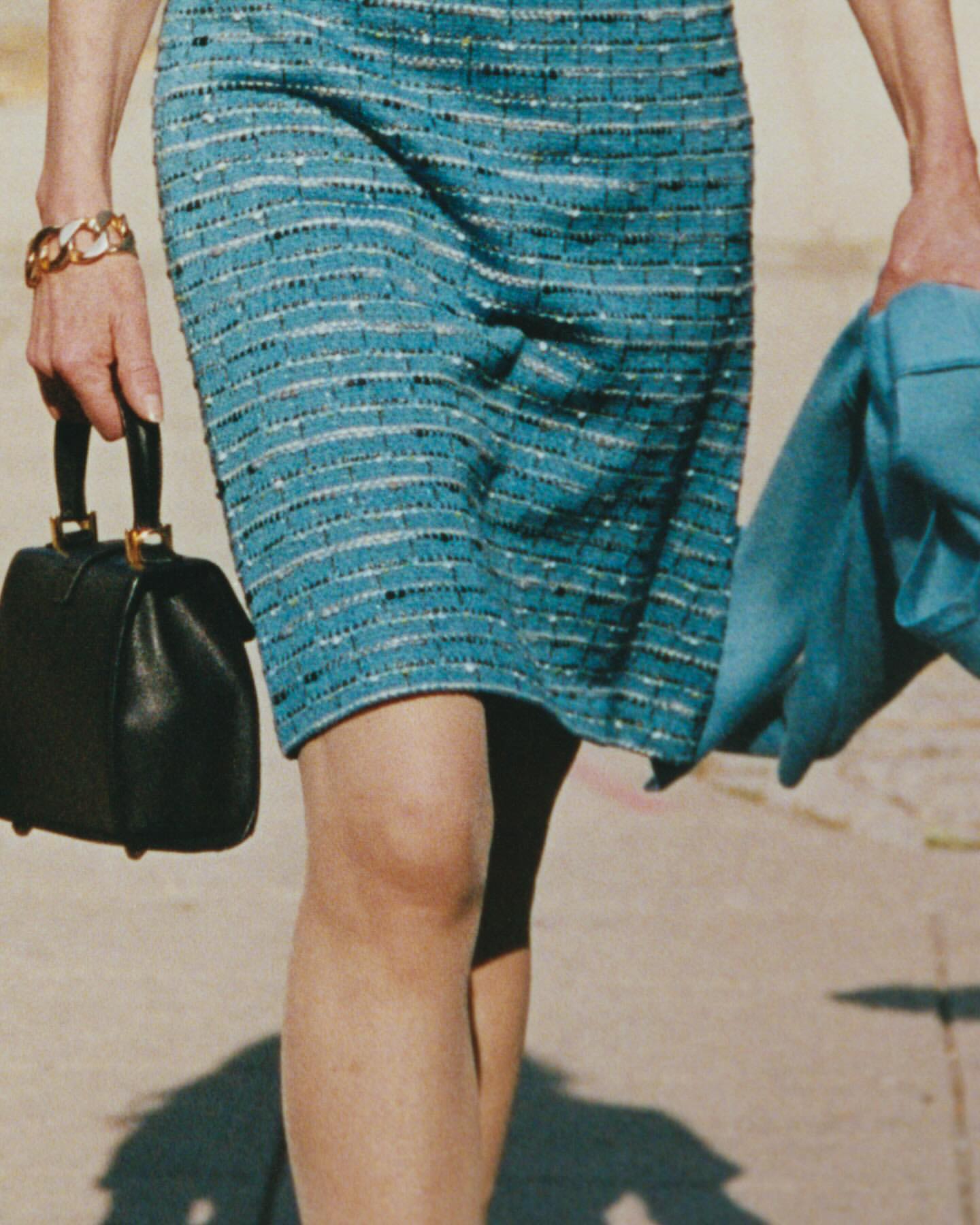 Close-up of a woman in a blue skirt holding a black purse and blue coat, capturing a fashionable, mid-stride moment in daytime.