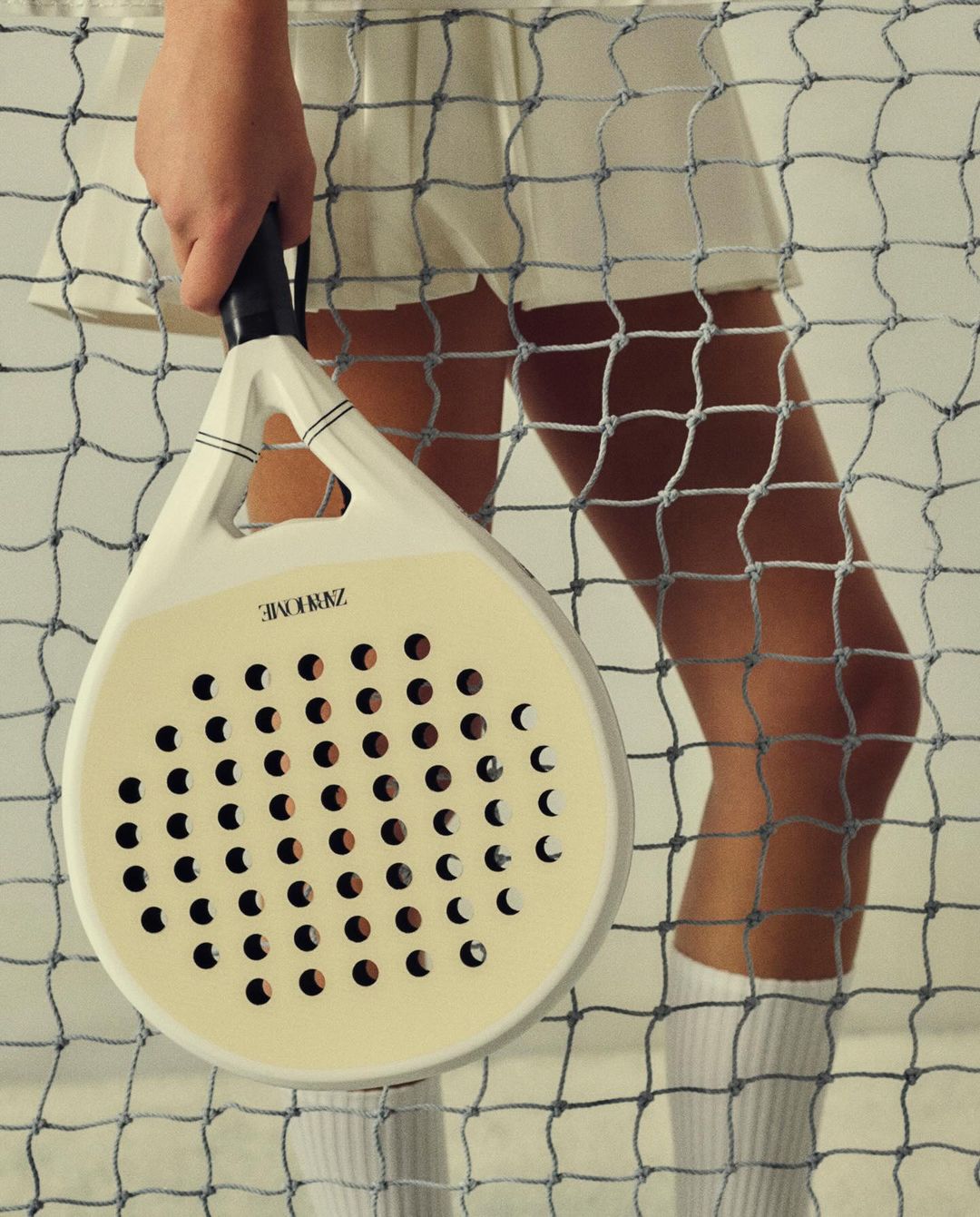 Woman holding a paddle racket in front of a tennis net, wearing a white skirt and knee socks, in a brightly lit indoor setting.