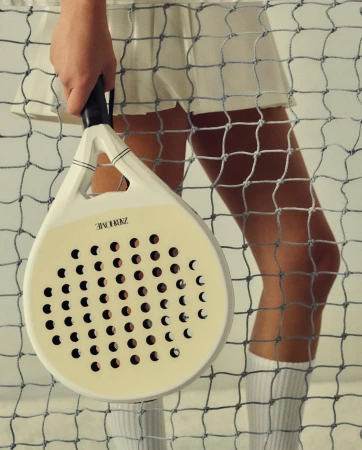 Woman holding a paddle racket in front of a tennis net, wearing a white skirt and knee socks, in a brightly lit indoor setting.