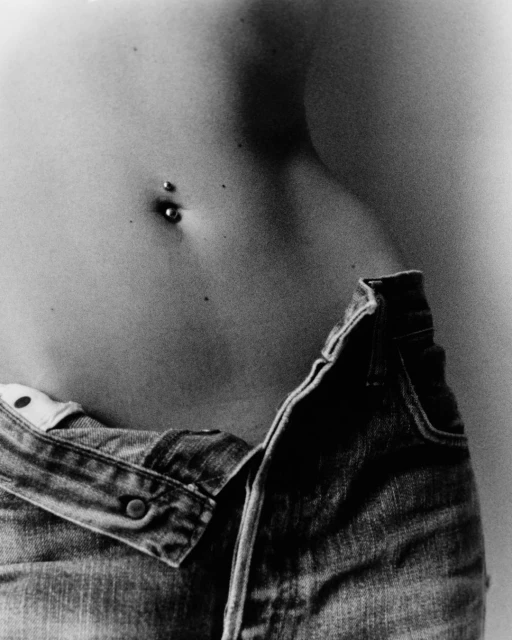 Close-up of a person's torso with a navel piercing, wearing low-rise denim jeans, captured in a black and white photograph.