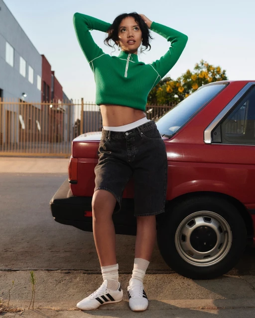 A young woman in a green sweater and black shorts leans against a red car in an urban setting with buildings and bright sky.