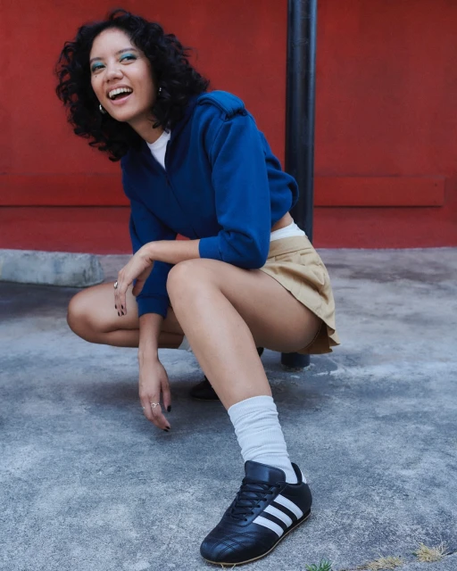 A smiling woman in a blue sweatshirt and tan shorts squats on concrete with black sneakers against a red wall in an eye-level shot.