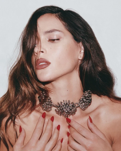 Adult woman poses in a close-up portrait in a photo studio with shoulder-length hair, dark lipstick, red nail polish, and a metal necklace.