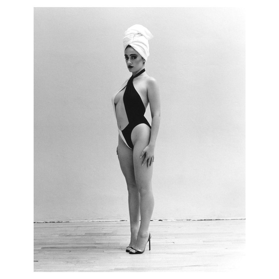 Adult woman posing in a black and white swimsuit and a white towel wrapped around her head, in a studio with a white wall.