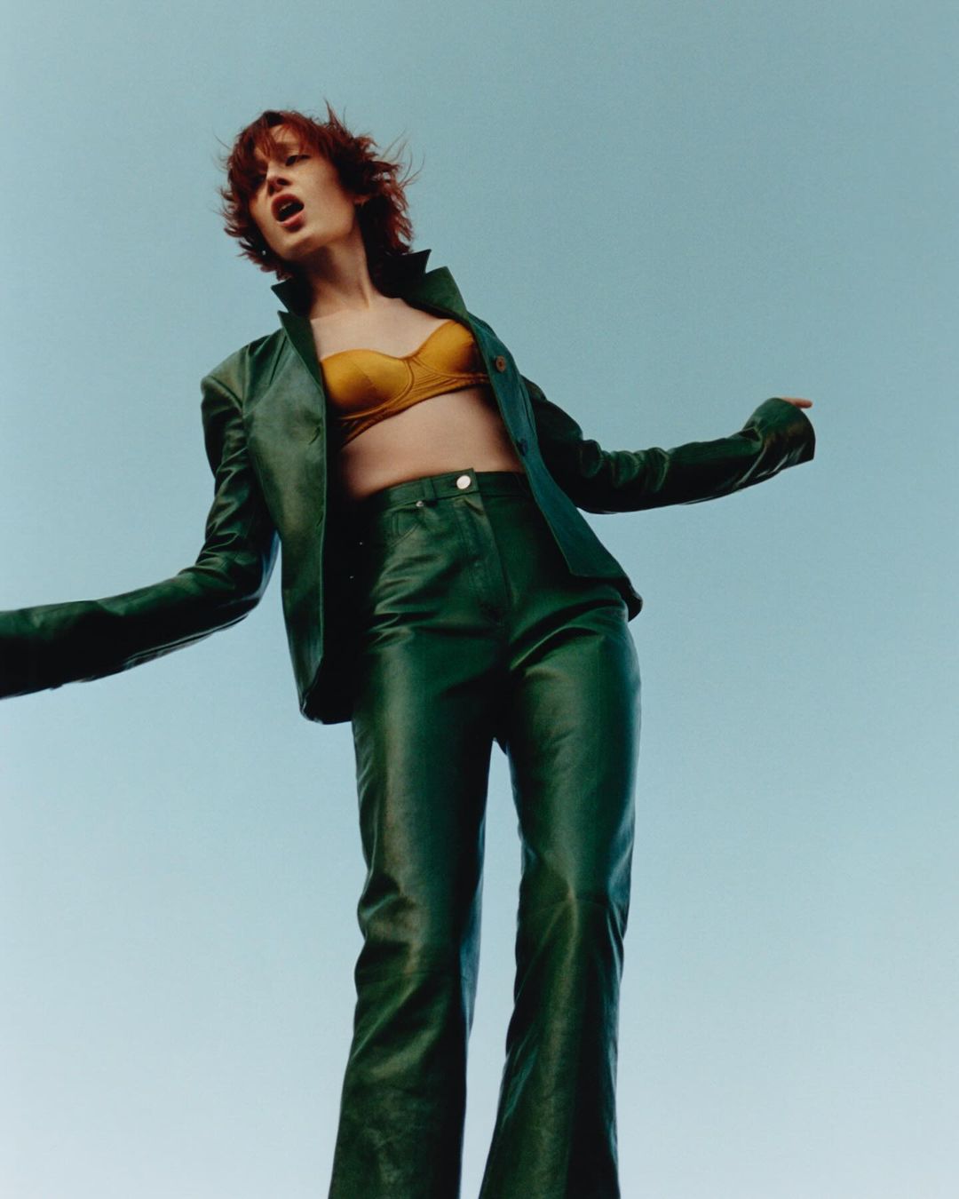 Woman in green leather jacket and pants stands against a blue sky, arms outstretched, creating a fashion-forward, confident, and dynamic visual.