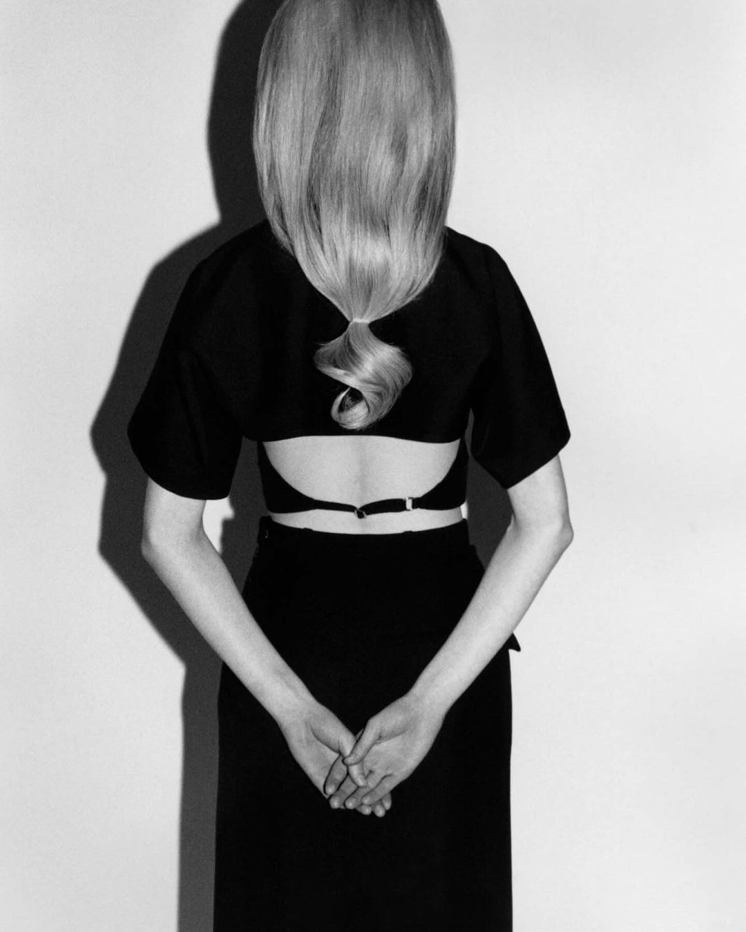 Back view of a blond woman in a black dress with an open back, her hands clasped behind her, set against a plain white background.