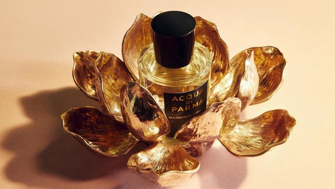 Still life shot of Acqua di Parma Magnolia Infinita perfume bottle in a golden lotus shaped holder against a pale background.