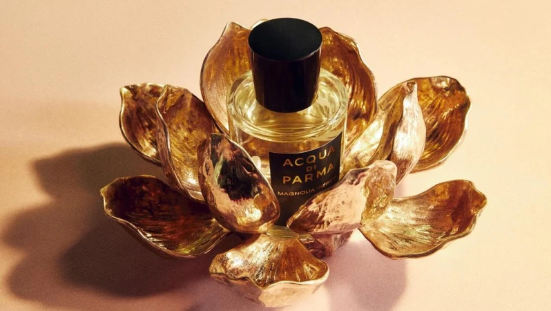 Still life shot of Acqua di Parma Magnolia Infinita perfume bottle in a golden lotus shaped holder against a pale background.
