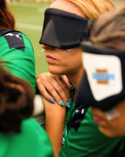 Close shot of three women in green sports jerseys wearing eyeshades with artificial nose plugs for goalball training on an indoor field.