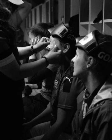 Close-up in black and white of a small group of people, one having makeup applied in an indoor locker room setting.