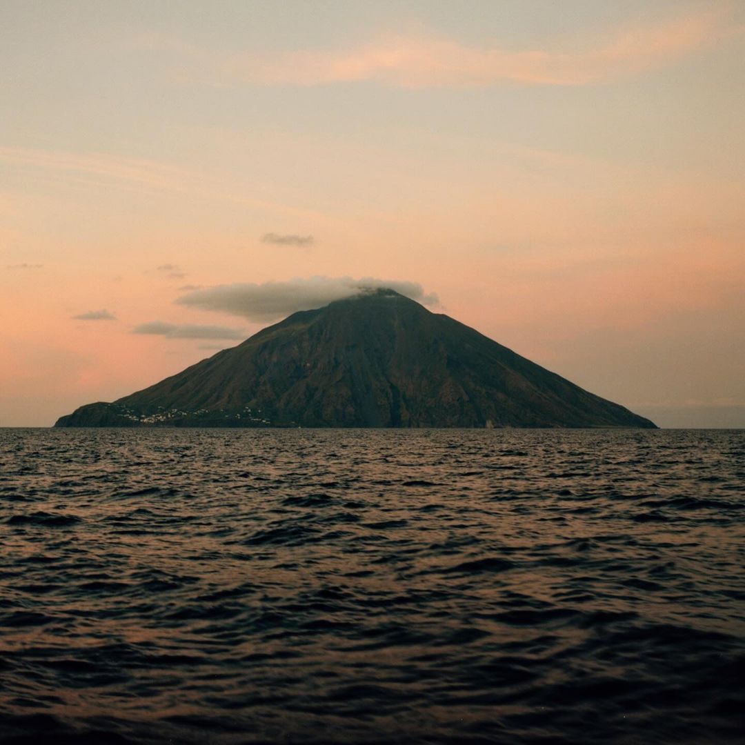 A volcanic island rises from the ocean under a pastel sunset sky with soft clouds, creating a serene and natural landscape.