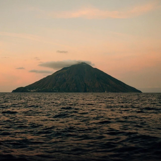 A volcanic island rises from the ocean under a pastel sunset sky with soft clouds, creating a serene and natural landscape.