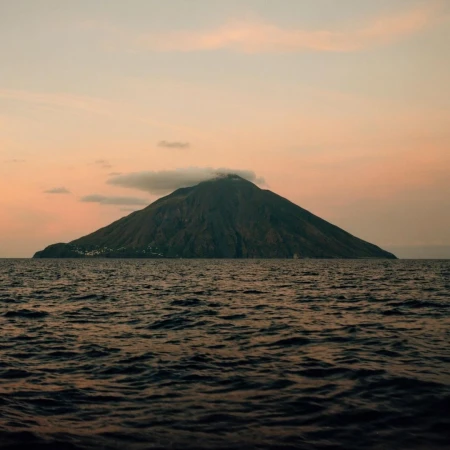 A volcanic island rises from the ocean under a pastel sunset sky with soft clouds, creating a serene and natural landscape.
