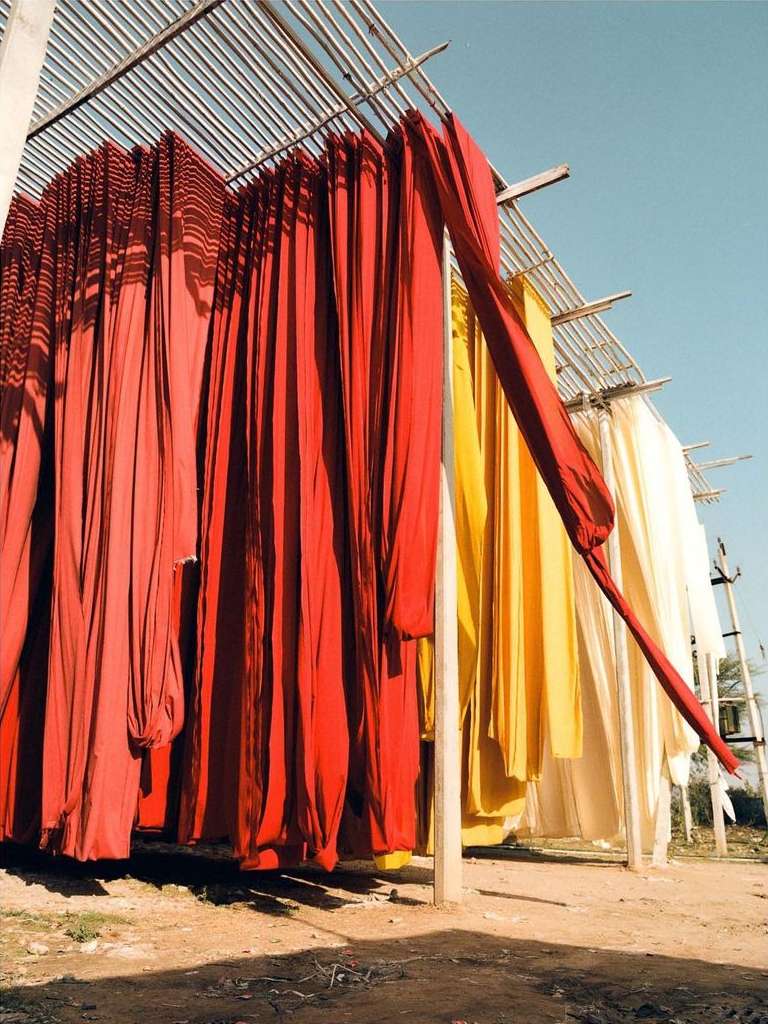 Outdoor scene featuring rows of dyed fabrics hanging to dry under a metal structure, displaying red, yellow, and cream hues.