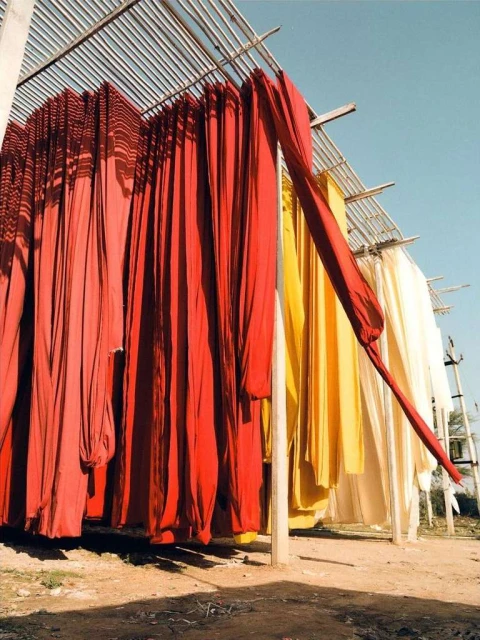 Outdoor scene featuring rows of dyed fabrics hanging to dry under a metal structure, displaying red, yellow, and cream hues.