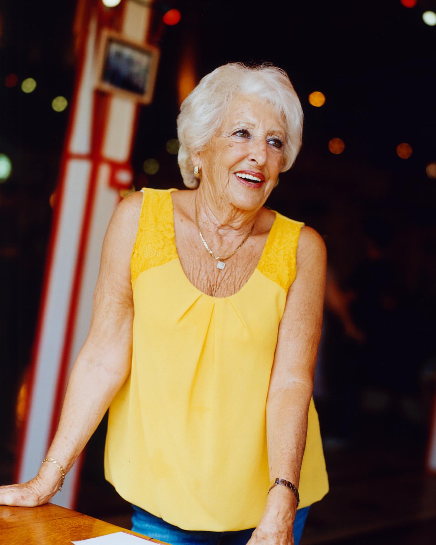 Smiling elderly woman with white hair stands indoors wearing yellow top and jewelry looking to the side in a warm, inviting atmosphere.