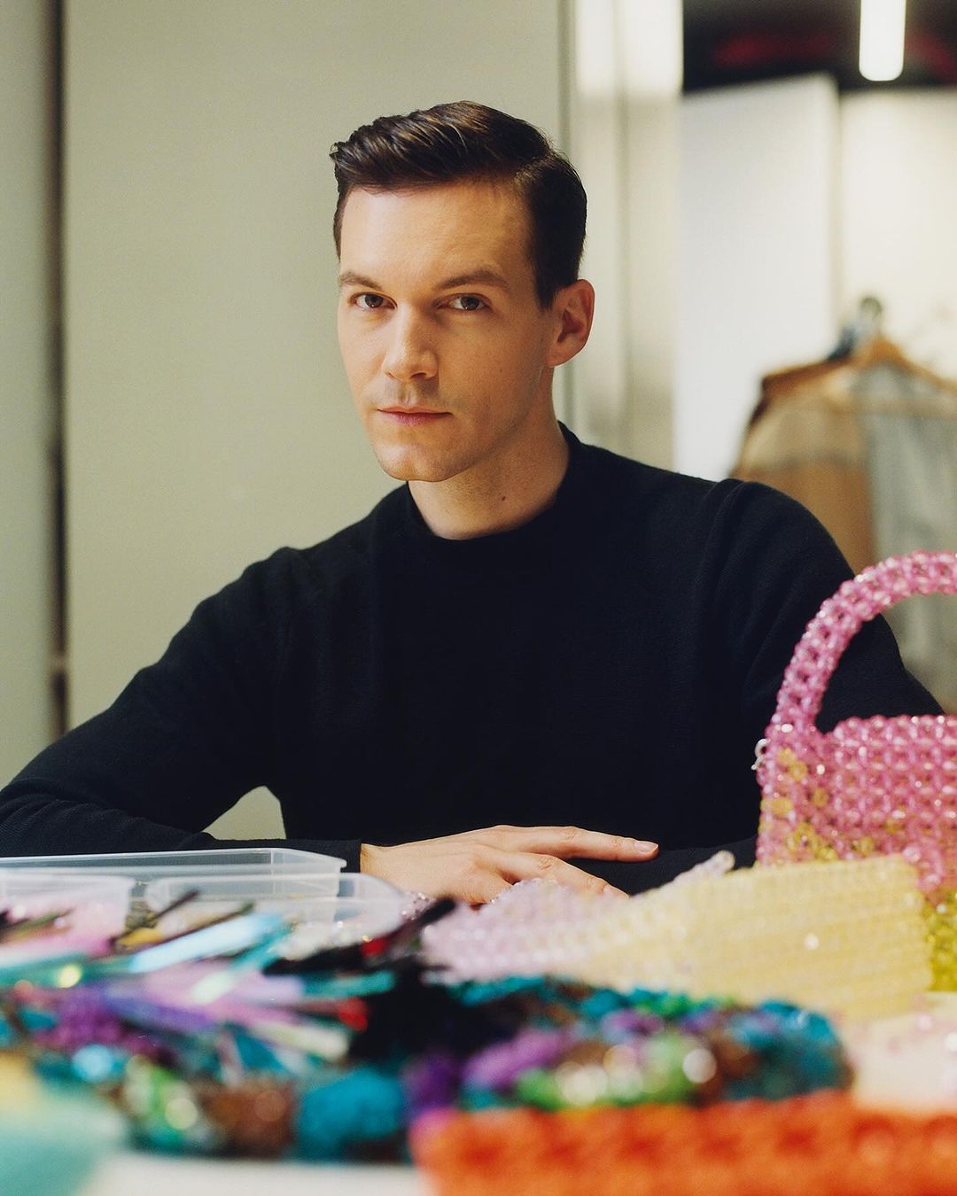 Portrait of a fair-skinned adult male with dark hair, seated at a table with beaded purses and craft supplies, inside a studio setting.