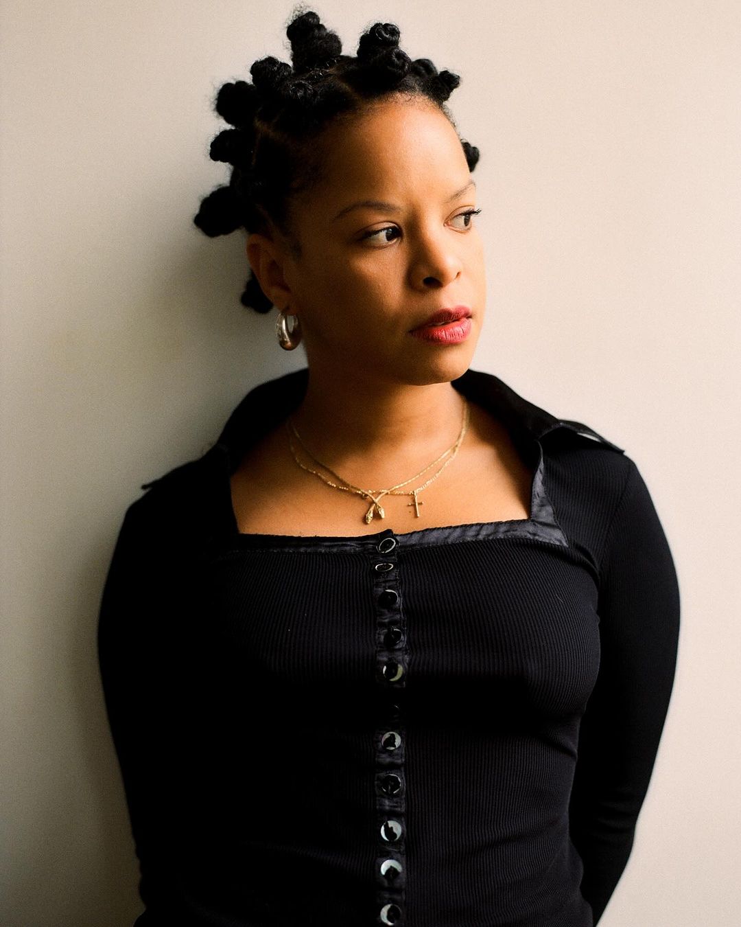 Portrait of a woman with knotted hair in a black top, featuring a neutral background and soft lighting. She wears gold necklaces.