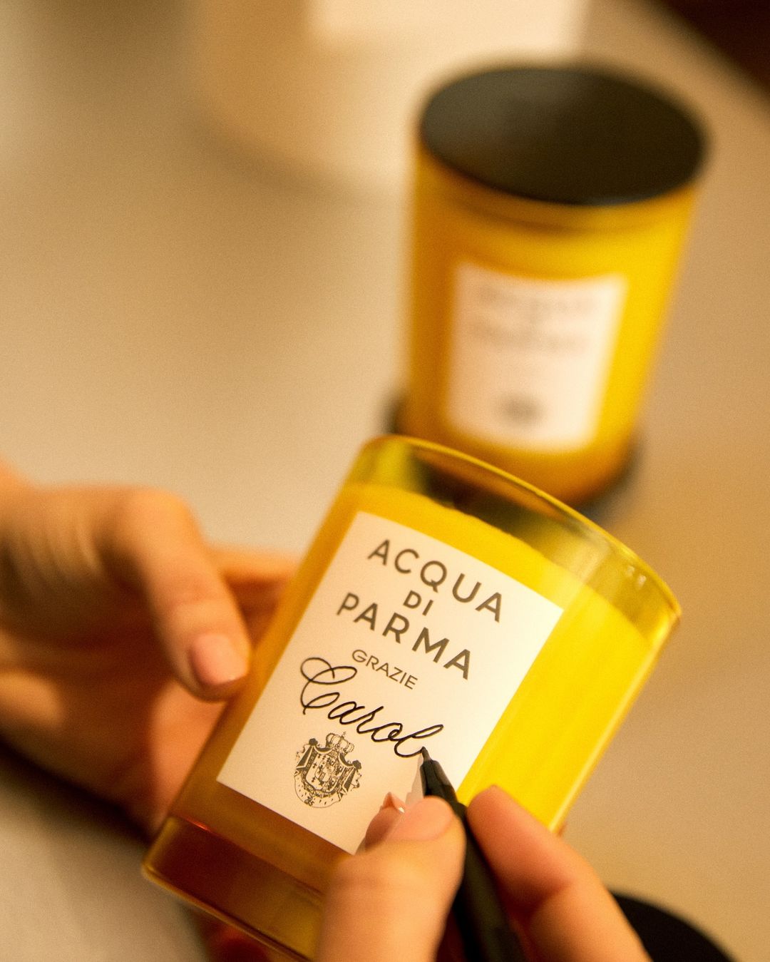 Close-up of a person holding a yellow Acqua di Parma candle while signing the label with a black marker, another candle behind.
