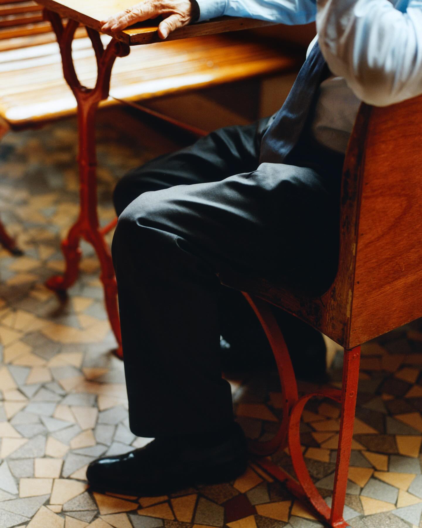Man seated at a table in a light blue shirt, dark pants, and shoes, resting his hand on the table, with a mosaic tile floor.