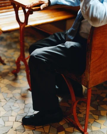 Man seated at a table in a light blue shirt, dark pants, and shoes, resting his hand on the table, with a mosaic tile floor.